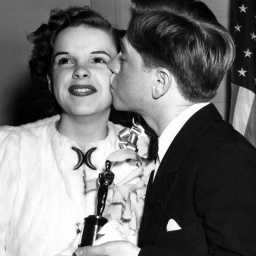 Eine junge Judy Garland hält eine Oscar Statue in der Hand und bekommen von einem jungen Mann einen Kuss auf die Backe