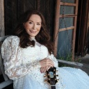 Countrylegende Loretta Lynn in einem weißen Kleid vor einem Holzhaus.
