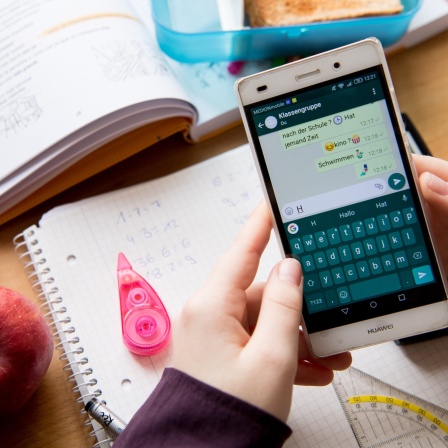Eine Schülerin sitzt mit ihrem Handy und ihren Schulsachen an einem Schreibtisch und hat einen Chat geöffnet.