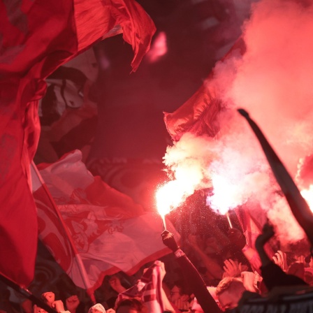 Nahaufnahme: In einer Fankurve brennen Leuchtfeuer und es wehen rote Fahnen.