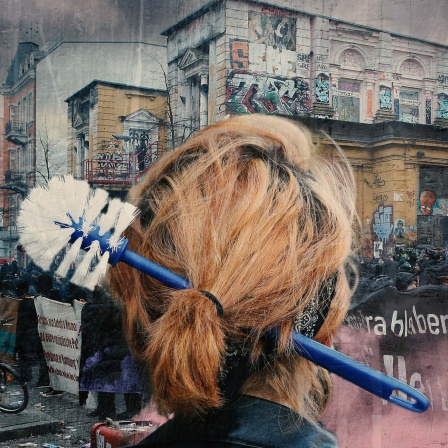 Person mit einer Klobürste im Haar, im Hintergrund Demonstranten vor einem besetzten Gebäude.