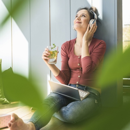 Eine weiße Frau hört mit Kopfhörern Musik, in der Hand ein gesundes Getränk, im Vordergrund unscharfe grüne Blätter