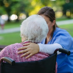 Eine Pflegeperson legt den Arm um eine alte Person. Die alte Person sitzt in einem Rollstuhl und ist lediglich von hinten zu erkennen.