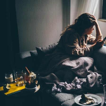 Eine junge Frau mit traurigem Gesichtsausdruck sitzt im Halbdunkel mit ihrem Smartphone in der Hand auf dem Sofa.