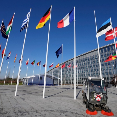 Ein leerer Fahnenmast steht zwischen den Nationalflaggen von Frankreich und Estland vor dem Nato-Hauptquartier. Eine Kehrmaschine säubert den Platz darunter.