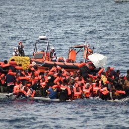 Ein Rettungsboot schwimmt hinter einem mit Migranten besetzten Boot.