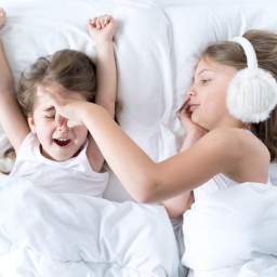 Ein kleines Mädchen liegt im Bett und schnarcht, seine Schwester trägt Kopfhörer und hält ihm die Nase zu