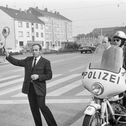 Siebziger Jahre, Schwarzweissfoto: An einem Zebrastreifen hebt ein Beamter eine Polizeikelle, daneben ein Polizist auf einem Motorrad.
