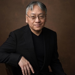 Kazuo Ishiguro sitzt vor neutralem Hintergrund und lächelt in die Kamera.