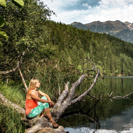 Eine Frau sitzt in lockerer Kleidung auf einem Baumstamm und schaut auf den Pigurber See im Naturpark Öztal 
