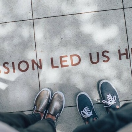Die Schuhe von zwei Personen sind zu sehen, die vor einem Schriftzug stehen, der am Boden angebracht wurde. Zu lesen sind die Worte: Passion brought us here.