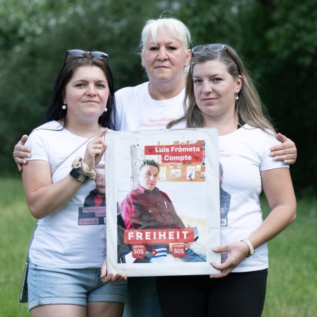 Maria (l) und Janie (r) Frometa Compte, Töchter des inhaftierten Deutsch-Kubaners Luis Frómeta Compte, und Silke Frometa Compte (M), Ehefrau, halten ein Foto von Luis Frometa Compte.