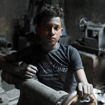 Ein Junge sitzt in einem dunklen Raum mit Werkzeugen und Maschinen; er blickt direkt in die Kamera; der 12-Jährige Hasibur Rahman lebt in Bangladesch und muss arbeiten (30.05.2020)