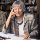 Dorothee Sölle, eine ältere Frau in einer blau-golden gestreiften Jacke, sitzt lächelnd an ihrem Schreibtisch, vor sich verschiedene Schriftstücke ausgebreitet.