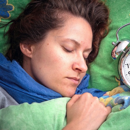 Eine Frau liegt im Bett und schläft, neben ihr liegt ein Wecker auf dem Kopfkissen.