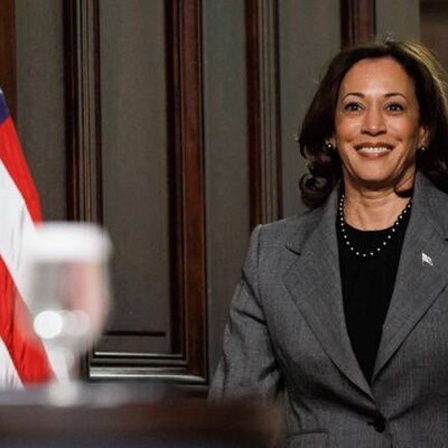 Kamala Harris betritt lächelnd einen Raum mit der US-Flagge auf der linken Seite.