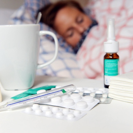 Eine Frau liegt im Hintergrund krank iim Bett, während im Vordergrund eine Teetasse, ein Fieberthermometer, Medikamente und Taschentücher neben einem Buch auf dem Nachttisch liegen.