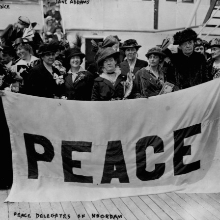 Historisches Schwarzweißfoto von Frauen, die 1915 auf einem Schiffsdeck stehen und ein Plakat mit der Aufschrift "Peace" in die Kameras halten.