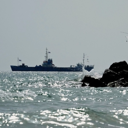 Zwei Männer angeln am Meer, während im Hintergrund ein mit 240 Tonnen Konserven und Hilfsgütern für den Gazastreifen beladenes Schiff in den Gewässern vor dem zypriotischen Hafen von Larnaca fährt.