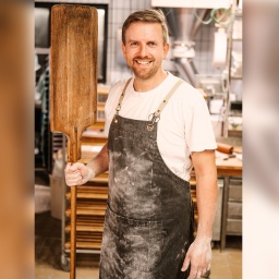 Ingmar Krimmer, Bäcker des Jahres 2024, über die Liebe zu Brot und Brezel. Er steht mit bemehlten Händen und Schürze in der Backstube und hält einen Brotschieber in der rechten Hand.