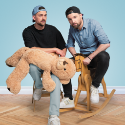 Nick und Leon  sitzen auf Kinderspielzeugen – der Mann links auf einem kleinen Stuhl mit einem großen Teddybären im Arm und der Mann rechts auf einem Holz-Schaukelpferd. Beide tragen legere Kleidung: der linke Mann ein schwarzes Shirt, eine graublaue Hose und eine umgekehrte Baseballkappe; der rechte Mann ein blaues Hemd, schwarze Jeans und eine schwarze Baseballkappe. Der Hintergrund ist in einem klaren, hellblauen Ton gehalten, während der Boden aus hellem Holzparkett besteht. 