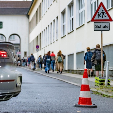 Pylonen stehen auf einer Straße vor einer Grundschule. Im Hintergrund gehen Schüler zum Schulgebäude. Im Vordergrund steht ein parkendes Auto mit rotem Bremslicht. 