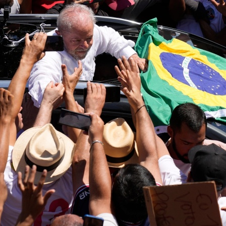Luiz Inácio Lula da Silva hält die Hand eines Unterstützers während er sich aus dem Auto lehnt. Auf dem Fahrzeug ist eine Brasilen-Flagge befestigt.