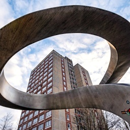 Durch eine geschlungene Skulptur hindurch ist ein Plattenbau zu sehen.