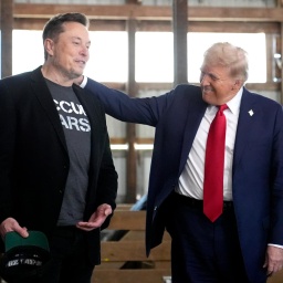 Donald Trump legt seine Hand aus die Schulter von Elon Musk und lächelt ihn an, während die beiden in einem Stall stehen.