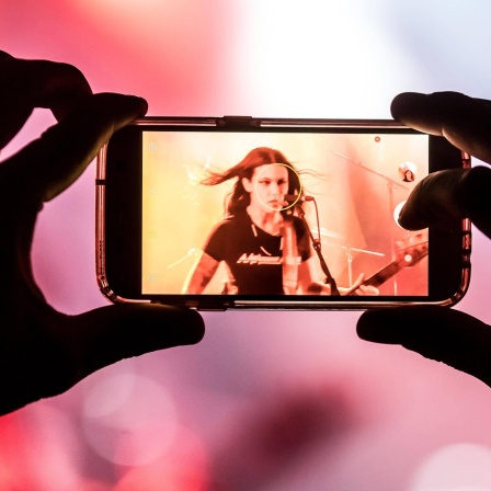 Ein Mensch filmt mit dem Handy eine Frau in einer Metalband auf einer Bühne.