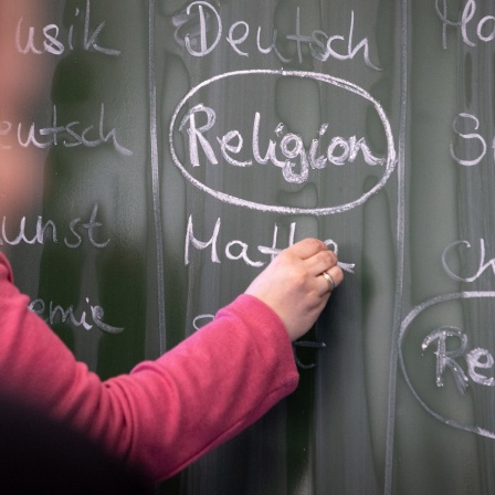 Eine Lehrerin schreibt mit Kreide einen Stundenplan mit Religionsunterricht an eine Tafel.