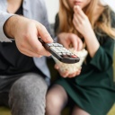 Zwei Menschen sitzen auf einer Couch mit einer Fernbedienung und Popcorn. 