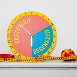 Uhr mit Anzeige "Fastenzeit/Essenszeit", daneben liegen ein Apfel und ein Maßband.
