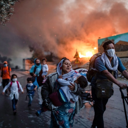 Ein brennendes Flüchtlingslager im Hintergrund. Davor fliehende Familien mit Kleinkindern.