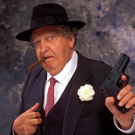 Ein Mafioso mit Hut und Zigarre hält eine Waffe aufrecht und schaut bedrohlich.