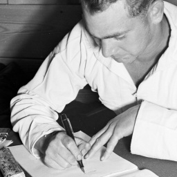 Ein Mann sitzt in weißem Overall an einem Tisch. Er hält einen Stift in der Hand und schreibt einen Brief.
