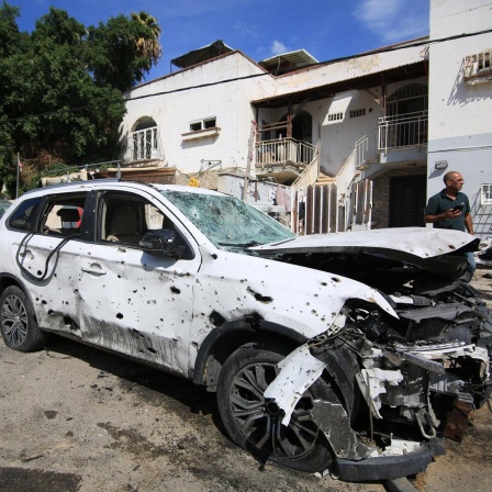 Im israelischen Ramla, eine Stadt zwischen Tel Aviv und Jerusalem, steht ein Auto, das durch Raketenbeschuss von Palästinensern demoliert ist. Rechts stehen Passanten.