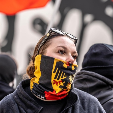 Eine junge Frau auf einer Demo verbirgt mit einem Schal mit dem Bundesadler auf schwarz-rot-goldenem Grund ihre untere Gesichtshälfte.