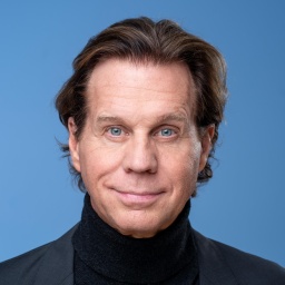 Schauspieler Thomas Heinze vor blauem Hintergrund mit schwarzem Jacket. Er schaut direkt in die Kamera