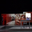 Bild der Bühne einer Aufführung von "Jugend ohne Gott" am Volkstheater Rostock. Im Hintergrund des Bühnenraums stehen Stellwände, die von einem übergroßen Gesicht bedeckt sind. Im Vordergrund Tische, am rechten Rand sitzt eine Frau im roten Kostüm auf einem Tisch und begutachtet die Szene. Am linken Bühnenrand steht ein Mann. 