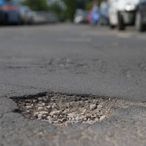 Mitten auf einer Straße ist ein Schlagloch zu sehen - im Hintergrund parken Autos am Straßenrand.