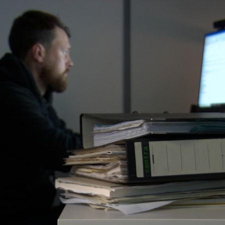 Ein Mann sitzt vor einem Computer, im Vordergrund liegt ein Aktenstapel