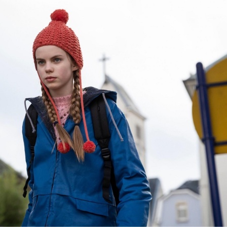Im Still aus "Mission Ulja Funk" ist Hauptdarstellerin Romy Lou Janinhoff mit einer roten Strickmütze zu sehen, hinter ihr verschwommen die Umrisse einer Kirche.