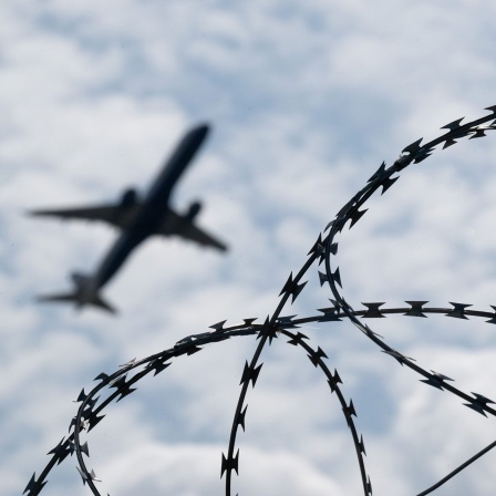 Ein Flugzeug hebt am Flughafen BER hinter einem Zaun mit Natodraht ab.