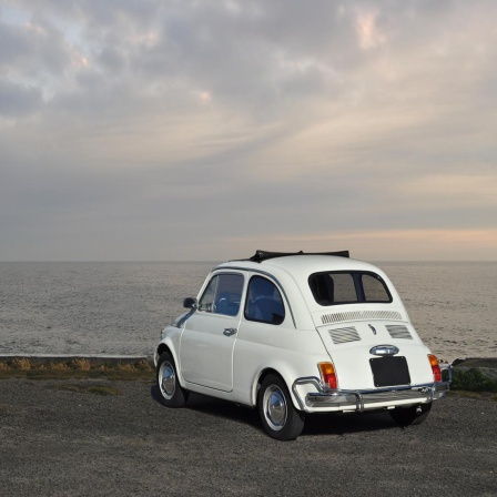 Ein alter, weißer Fiat steht am Meer, der Fahrer sitzt in dem Auto und blickt in Richtung Meer.