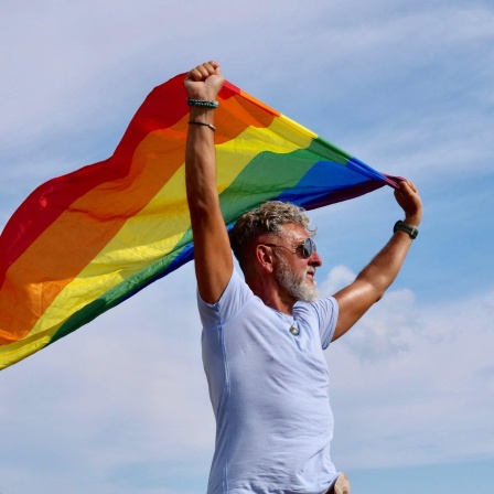Portrait senior of a gray-haired elderly Caucasian man with a beard and sunglasses holding a rainbow LGBTQIA flag against a sky
