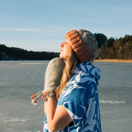 Eine Frau im Winter mit Mütze, Handschuhen und kurzen Ärmeln, die in die Sonne schaut, neben ihr ein See.