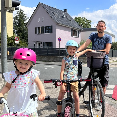 Ein Vater steht mit seinen zwei Kindern an der Kreuzung. Sie sind mit Fahrrädern unterwegs.