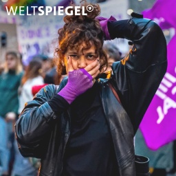 Eine Frau schaut in die Kamera, hält sich eine Hand an den Mund, die andere an den Hinterkopf, trägt lila Handschuhe