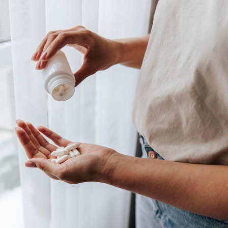 Eine Frau steht am Fenster und schüttet weiße Pillen aus einer weißen Dose in ihre Hand.